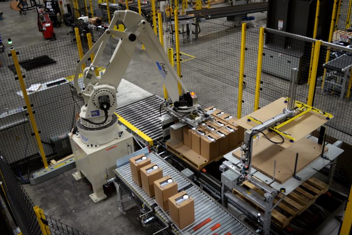 The picture shows a white Columbia Okura palletizer placing small crates on a conveyor belt.