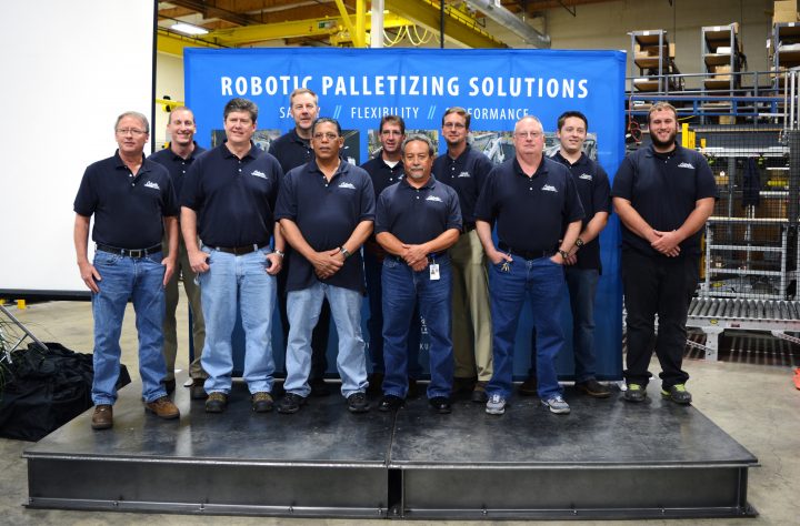 11 men wearing Columbia polos stand in front of the blue Robotic Palletizing Solutions banner. They look happy and proud.
