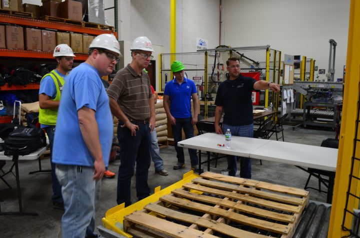 5 men wearing white safety helmets are looking at the pallet. One is pointing at something behind the camera